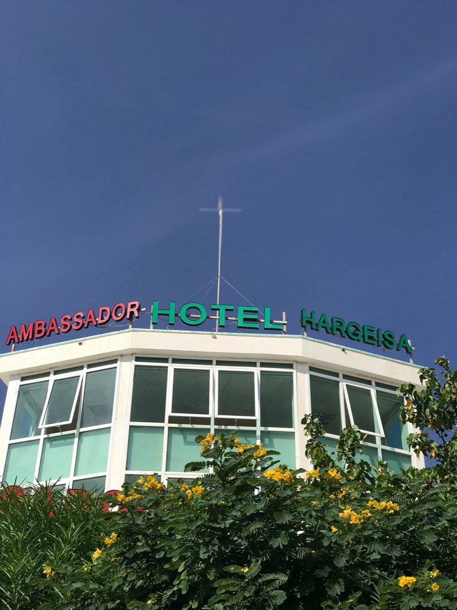 The Ambassador Hotel Hargeisa sign on the rooftop of its glass-fronted tower, above flowering trees
