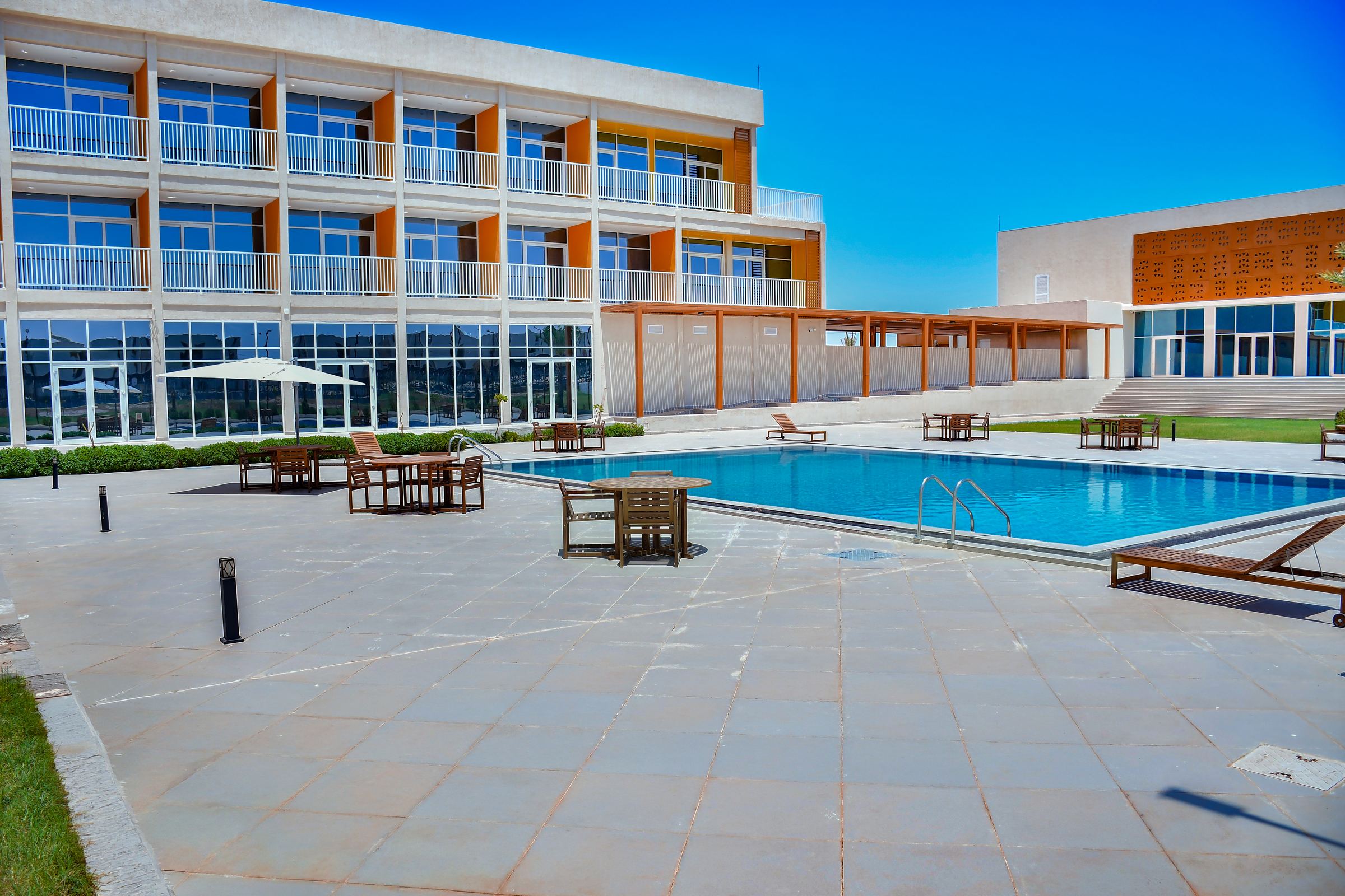 The outdoor pool with tables and sun loungers, in front of rooms with balconies